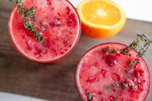 Two glasses of raspberry cocktail garnished with thyme sprigs on a wooden surface with half an orange