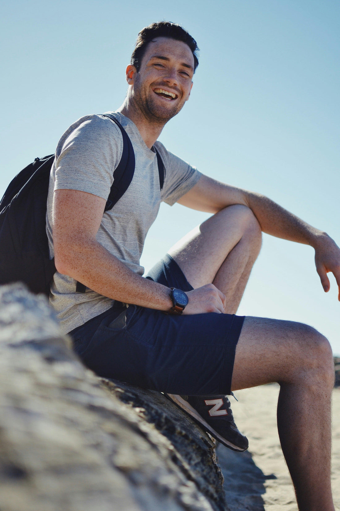 Smiling man in gray shirt and shorts sitting on rock with backpack under clear blue sky