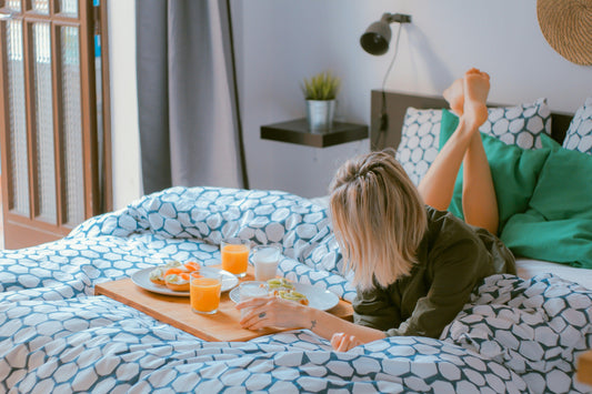 Woman lying on bed with patterned bedding enjoying breakfast tray with juice and fruit