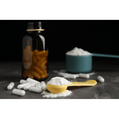 Yellow scoop with white powder and white capsules near amber bottle and blue scoop on dark surface