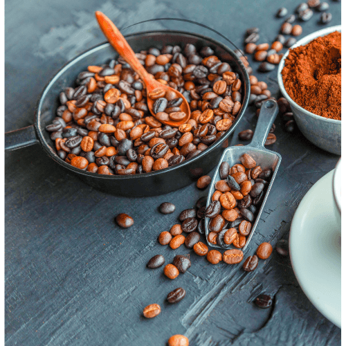 Roasted coffee beans in a pan with wooden spoon, metal scoop, ground coffee in bowl on dark textured surface