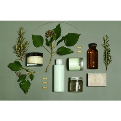 Natural personal care items including green leaves, herb sprigs, cream jars, lotion bottle, brown glass bottle, soap bar, and capsules on green background