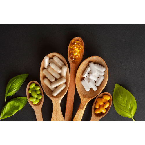 Wooden spoons with various vitamins and supplements including capsules, tablets, and soft gels on black background with green leaves