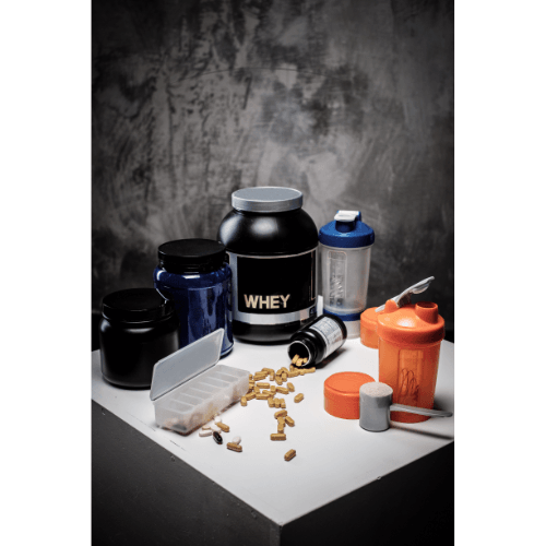 Whey protein container, supplement jars, capsules, and two shaker bottles on a white surface